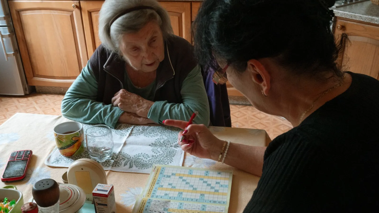 Une aide à domicile aide une dame âgée à remplir une grille de mots croisés à la table de sa cuisine
