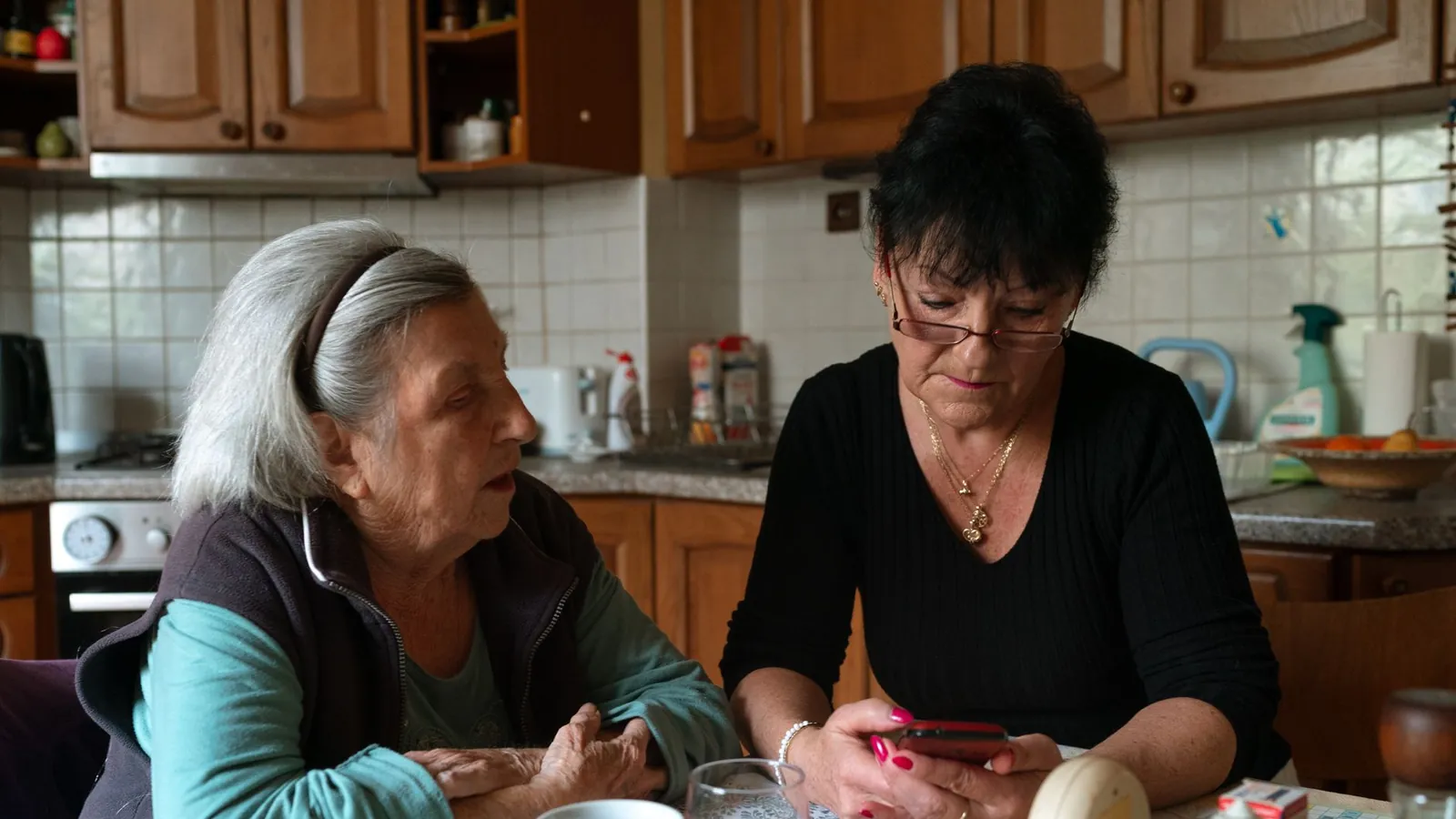 Une aide à domicile et une dame âgée discutent à la table de la cuisine, un téléphone entre les mains