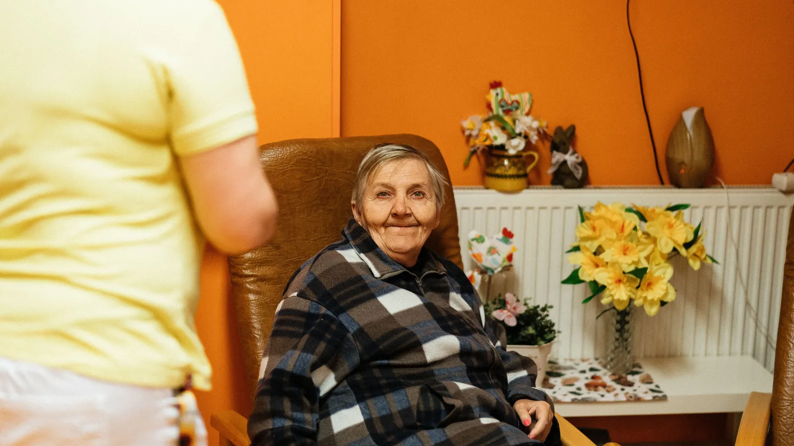 Une femme âgée sourit, assise dans un fauteuil de sa chambre en maison de retraite, avec un bouquet de jonquilles derrière elle