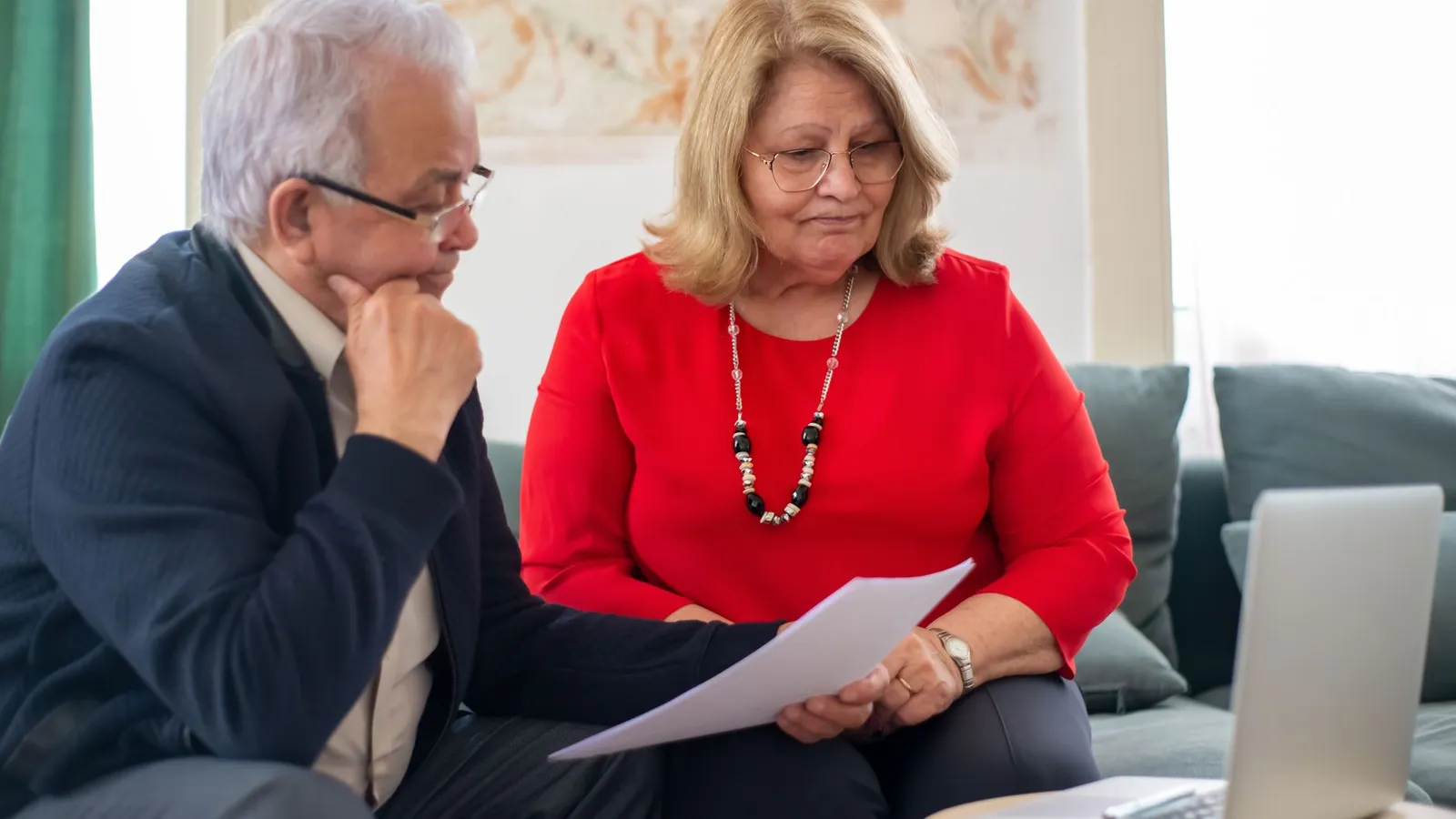 Un couple de seniors examine ensemble des documents sur un canapé, un ordinateur portable devant eux