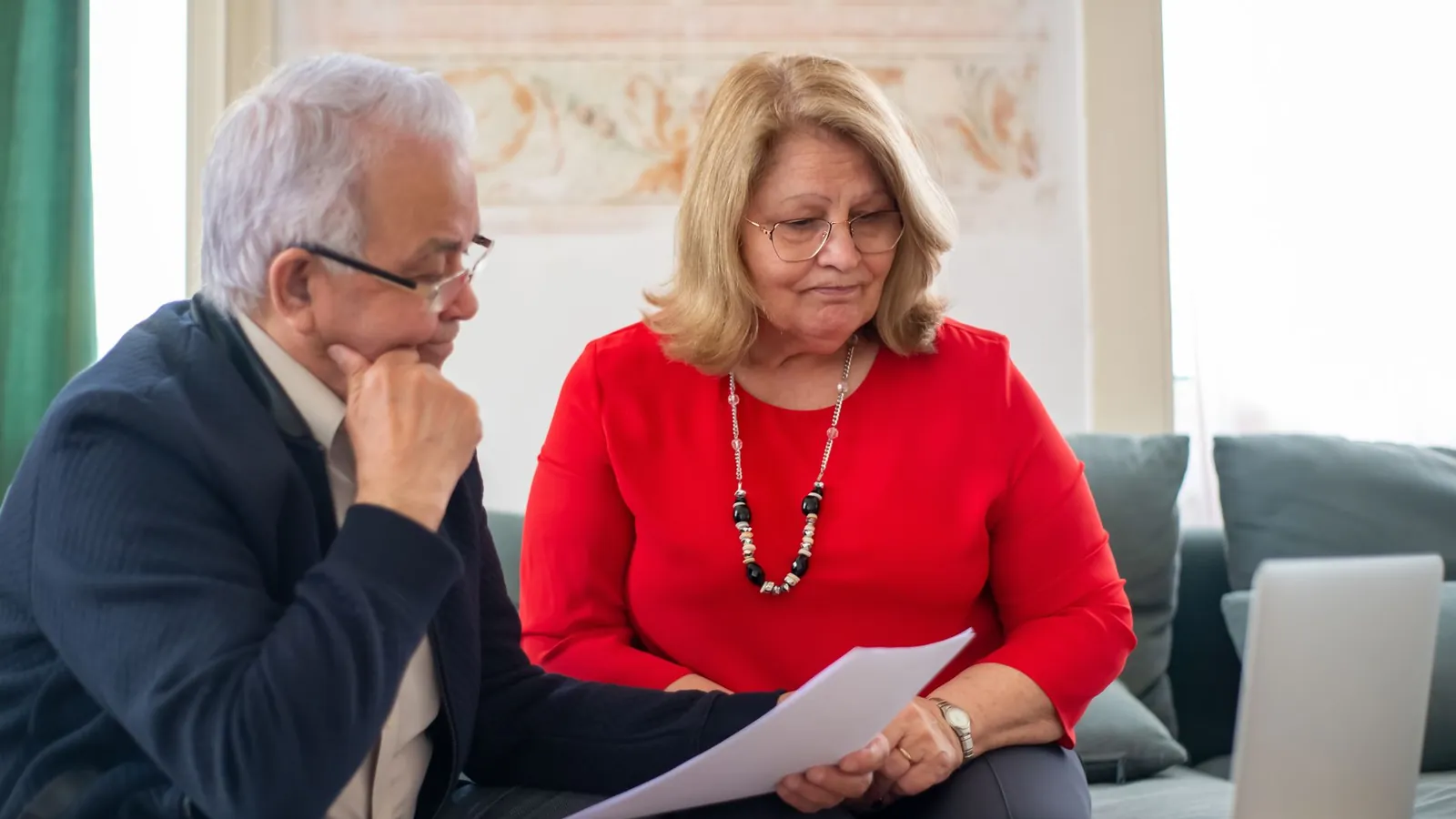 Un couple de seniors regarde ensemble un document papier, un ordinateur portable posé près d’eux