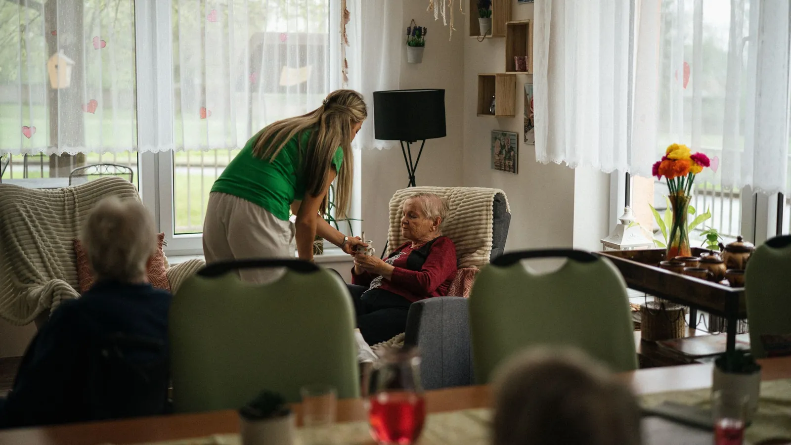 Une aide-soignante se penche vers une résidente installée dans un fauteuil, dans la salle commune lumineuse d’un établissement pour personnes âgées
